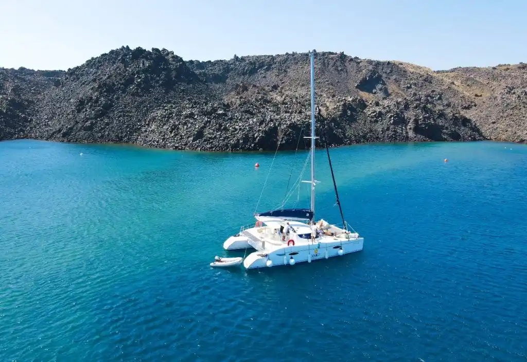 Καταμαράν αγκυροβολημένο σε τιρκουάζ νερά, κάτω από σκούρα βραχώδη ακτή