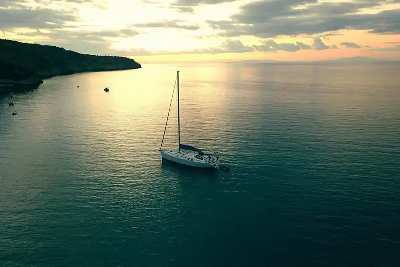 Sailing yacht on a calm sea at sunset, seen from above, a wooded headland on the left