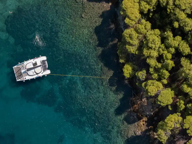 Catamaran moored with a line to the pine-covered shore at Agia Kyriaki, seen from above