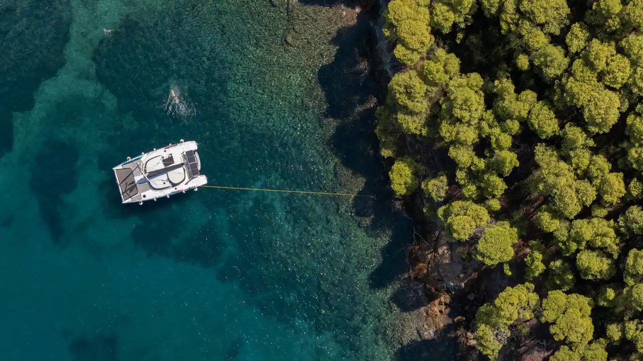 Catamaran moored with a line to the pine-covered shore at Agia Kyriaki, seen from above