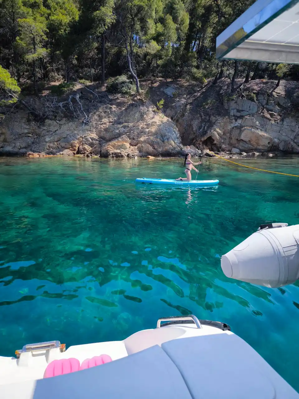 Paddleboarder in turquoise water below pine-covered rocks, seen from a catamaran