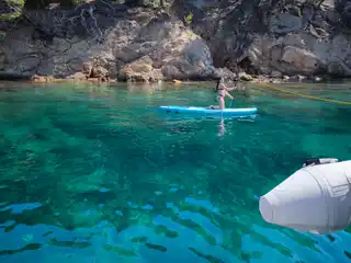 Paddleboarder in turquoise water below pine-covered rocks, seen from a catamaran