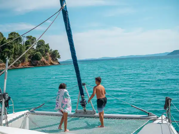 Two children on a catamaran’s net looking toward a pine-covered headland
