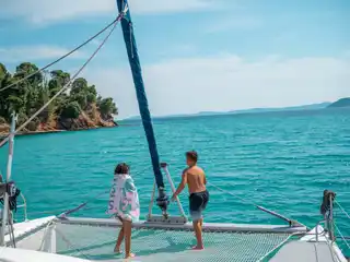 Two children on a catamaran’s net looking toward a pine-covered headland
