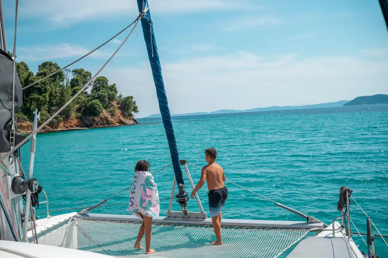 Two children on a catamaran’s net looking toward a pine-covered headland