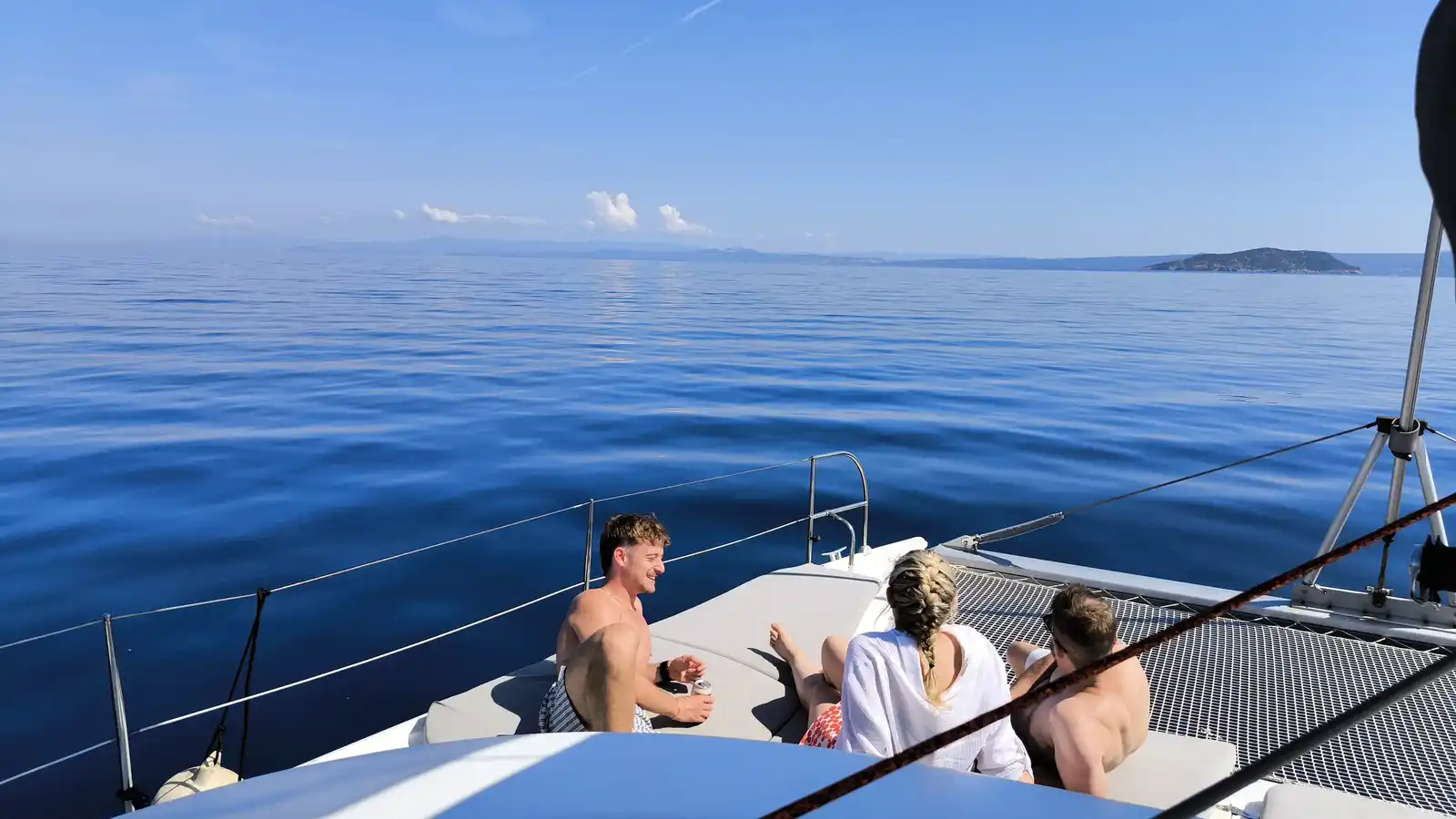Friends on a catamaran’s bow on a calm open sea, an islet in the distance