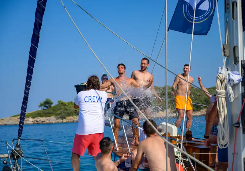 Crew member splashing laughing guests with a bucket of water on a sailing yacht