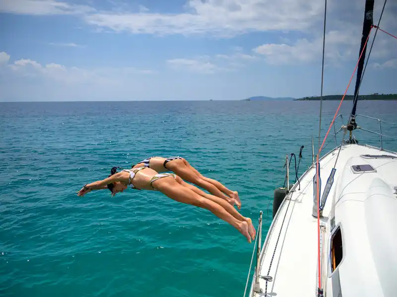 Two women diving together from a sailing yacht’s bow into turquoise water