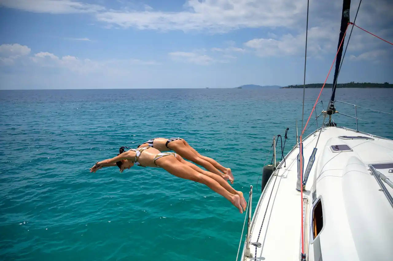 Two women diving together from a sailing yacht’s bow into turquoise water