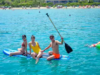 Three friends sitting on a paddleboard in turquoise water off a sandy beach