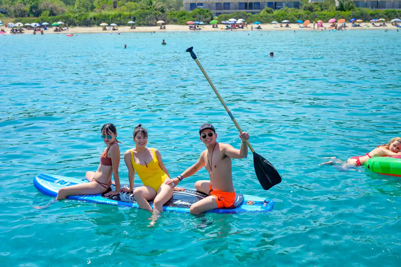 Three friends sitting on a paddleboard in turquoise water off a sandy beach