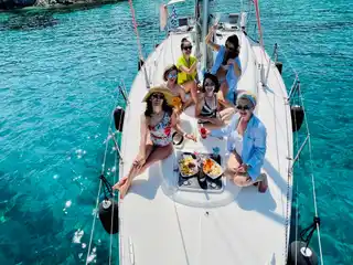 Friends on the bow of a sailing yacht in a turquoise cove below pines