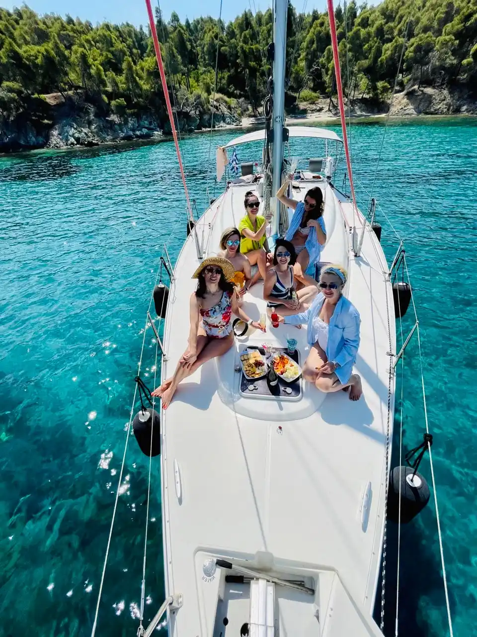 Friends on the bow of a sailing yacht in a turquoise cove below pines