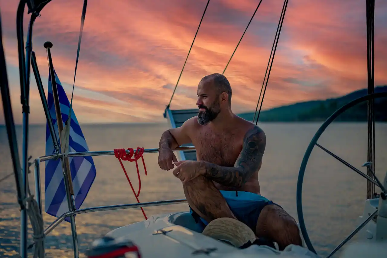 Man sitting at a sailing yacht’s helm under a red evening sky