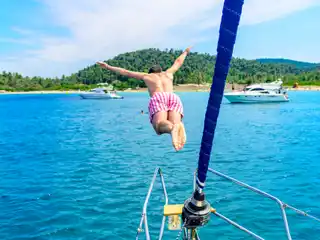Man diving from a sailing yacht’s bow toward a wooded beach