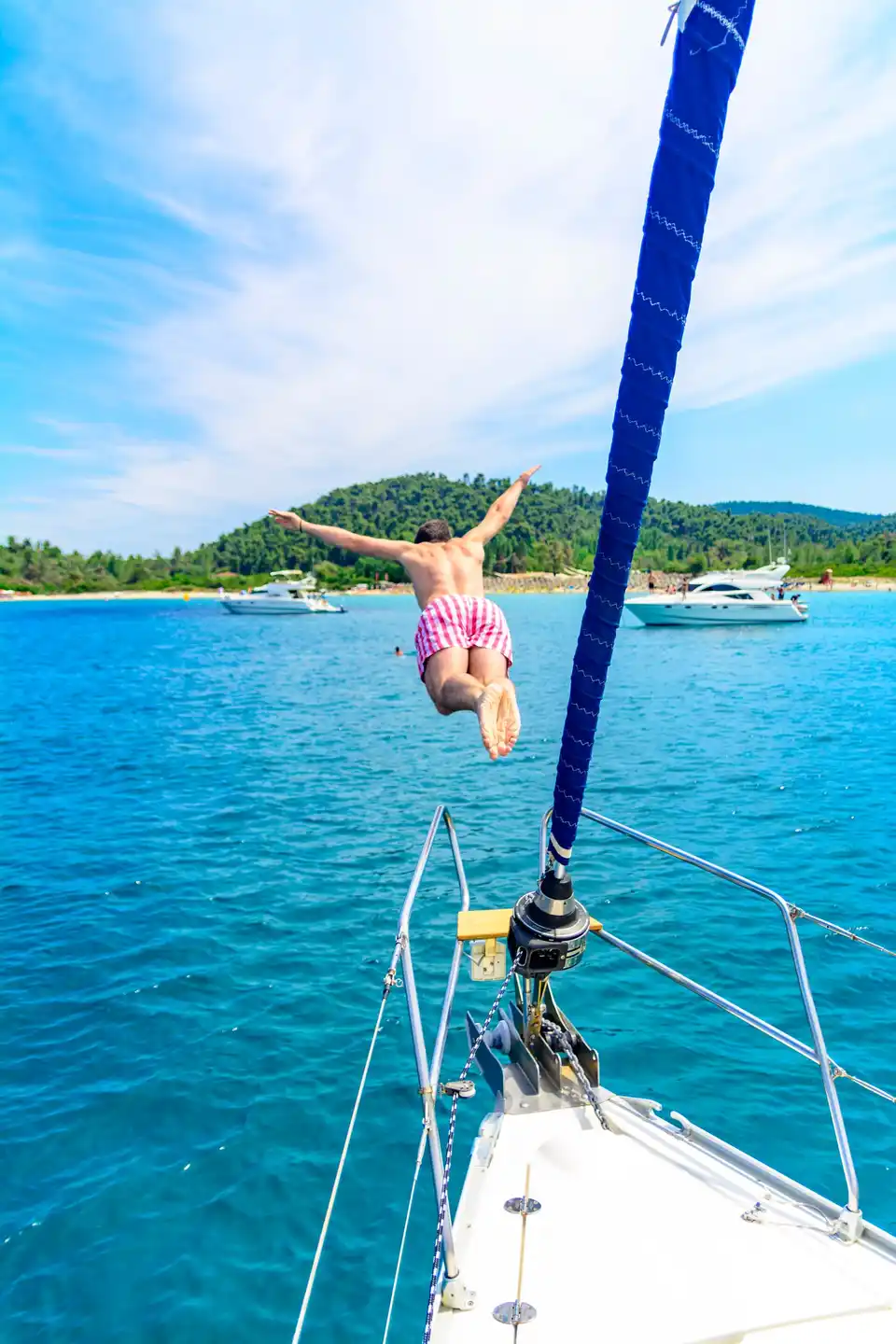Man diving from a sailing yacht’s bow toward a wooded beach
