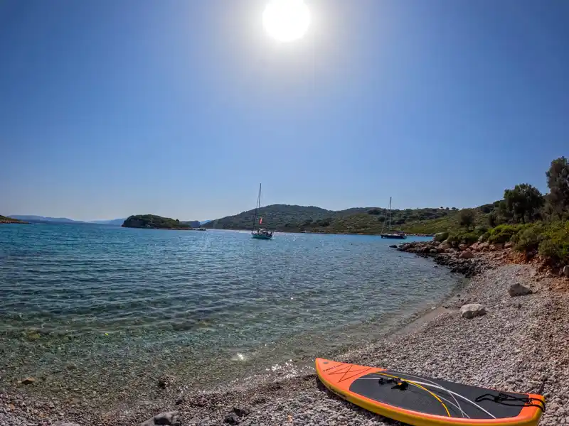 Πορτοκαλί σανίδα SUP σε παραλία με βότσαλα, με ιστιοπλοϊκά στην άγκυρα στον όρμο