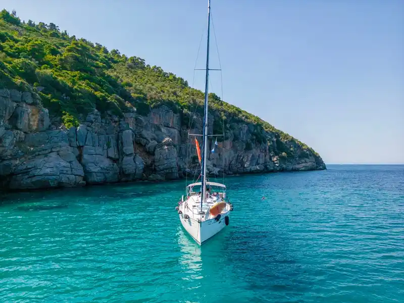 Sailing yacht anchored in turquoise water below a pine-topped limestone cliff off Paliouri