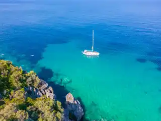 Sailing yacht anchored in turquoise water off a rocky, pine-covered point