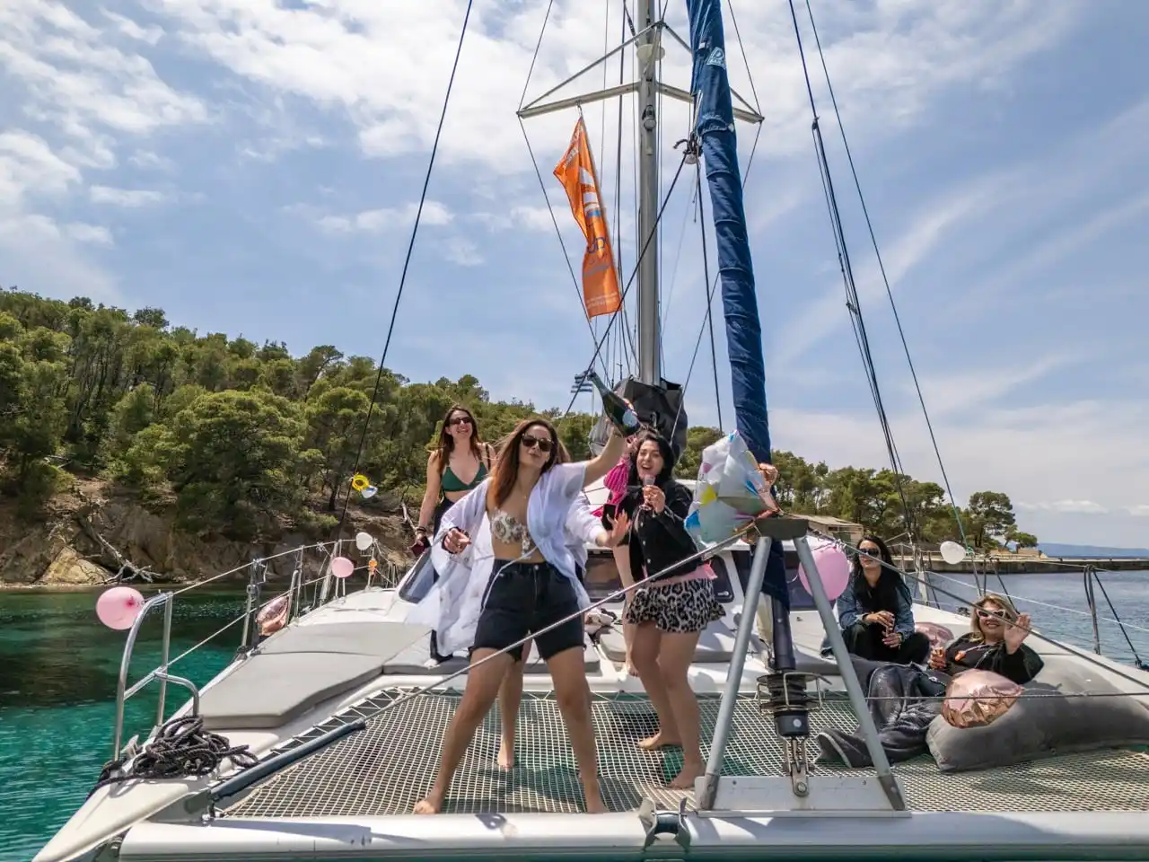 Friends celebrating with balloons on the bow of a catamaran near a pine shore