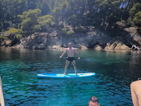 Επιβάτης όρθιος σε σανίδα SUP δίπλα σε βραχώδη ακτή με πεύκα, κοντά στο Πόρτο Καρράς