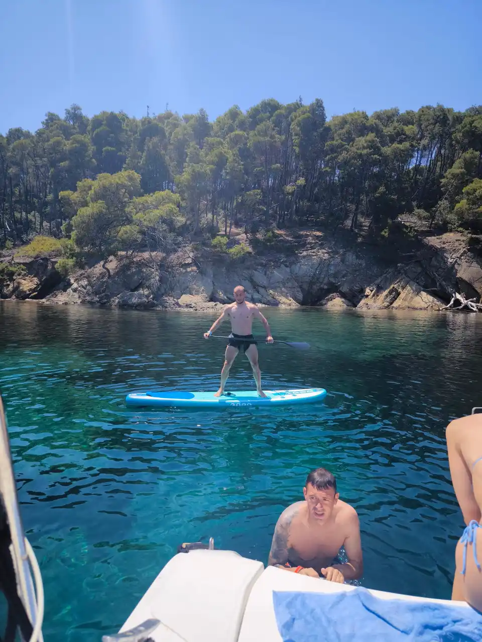Επιβάτης όρθιος σε σανίδα SUP δίπλα σε βραχώδη ακτή με πεύκα, κοντά στο Πόρτο Καρράς