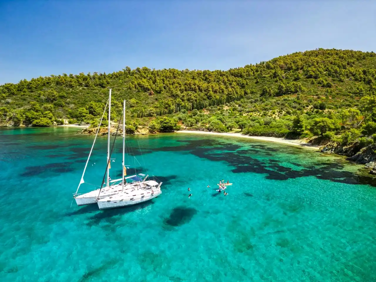 Two sailing yachts anchored in turquoise water below a pine-covered hill near Porto Carras
