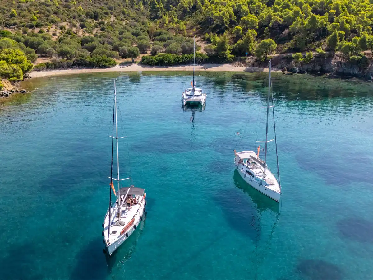 Δύο ιστιοπλοϊκά κι ένα καταμαράν σε τιρκουάζ όρμο κάτω από πεύκα, κοντά στο Πόρτο Καρράς