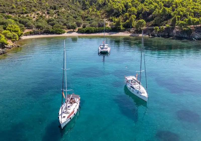 Δύο ιστιοπλοϊκά κι ένα καταμαράν σε τιρκουάζ όρμο κάτω από πεύκα, κοντά στο Πόρτο Καρράς