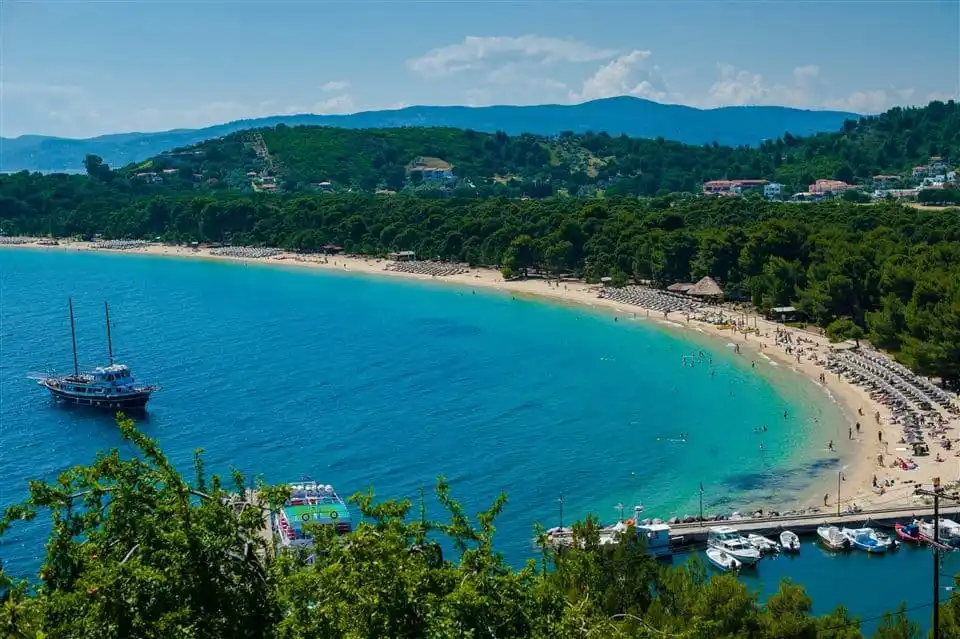 Long sandy beach lined with pines, a wooden two-masted boat at anchor in the bay
