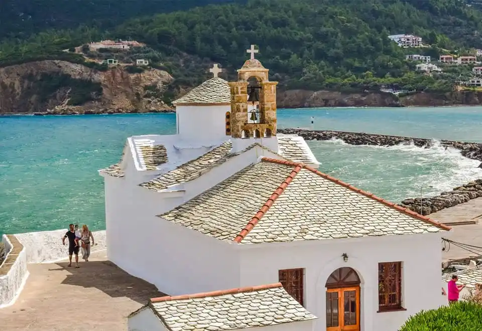 White chapel with a stone roof and bell tower by a harbour, green hills across the water