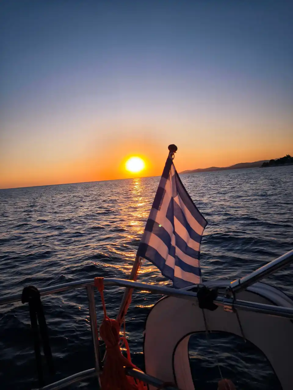 Sun setting over the sea behind a Greek flag on a yacht’s stern