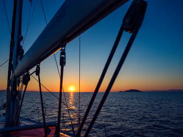 Sun setting over the sea behind a sailing yacht’s boom and rigging
