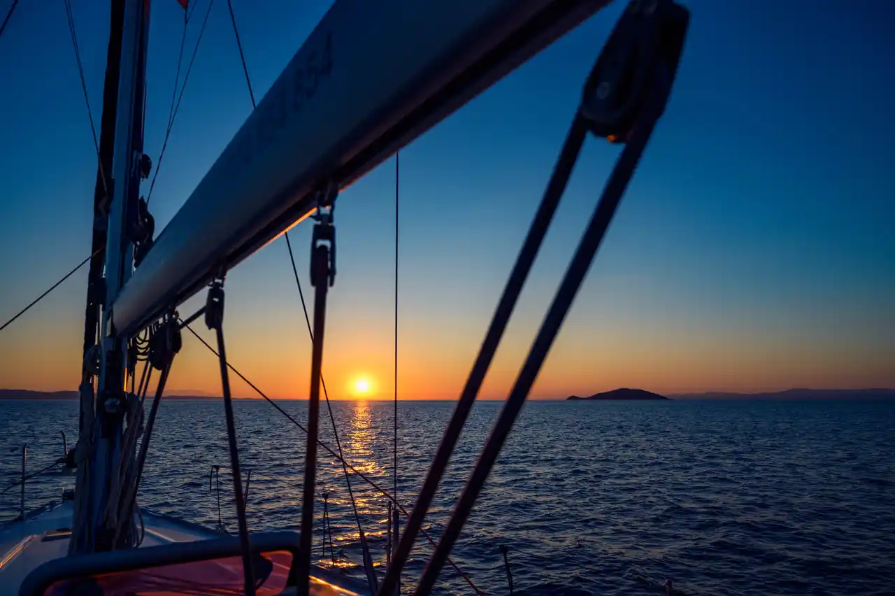 Sun setting over the sea behind a sailing yacht’s boom and rigging