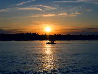 Small boat crossing a calm sea as the sun sets behind a wooded shore