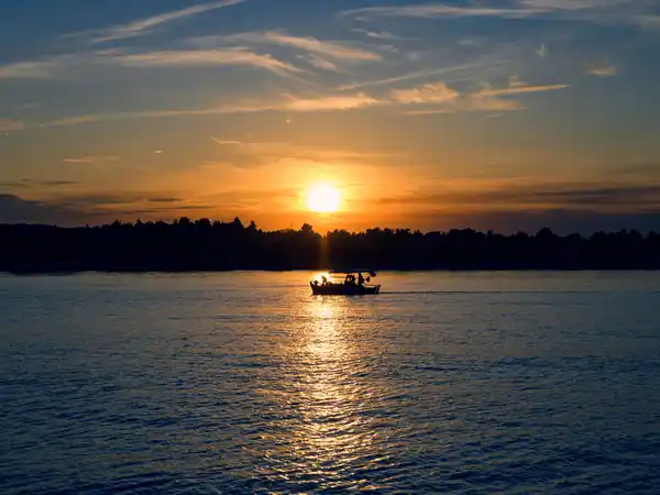 Small boat crossing a calm sea as the sun sets behind a wooded shore