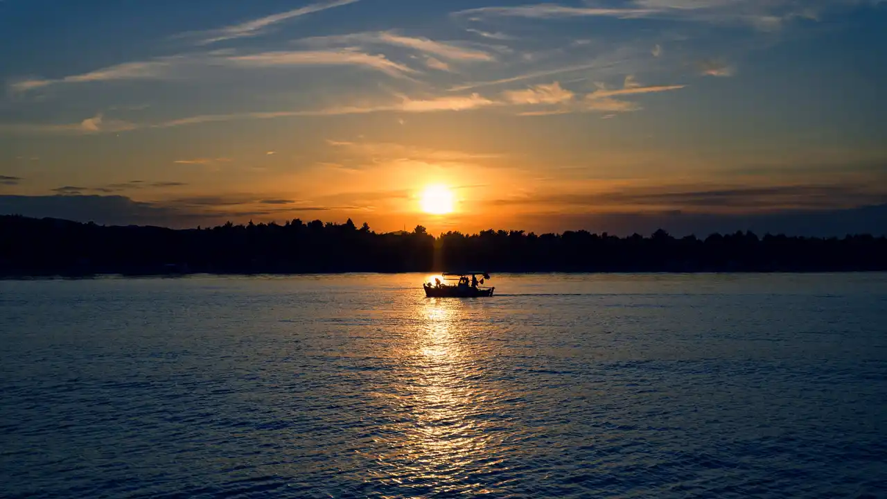 Small boat crossing a calm sea as the sun sets behind a wooded shore