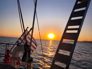 Sun setting over the sea, seen past a yacht’s swim ladder and Greek flag