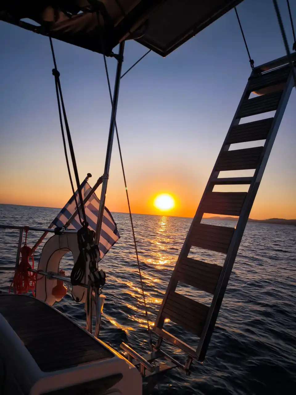 Sun setting over the sea, seen past a yacht’s swim ladder and Greek flag