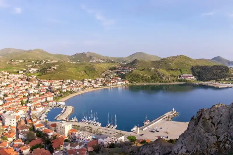 Πόλη με κόκκινες στέγες γύρω από λιμάνι με ιστιοπλοϊκά, και πράσινοι λόφοι από πίσω