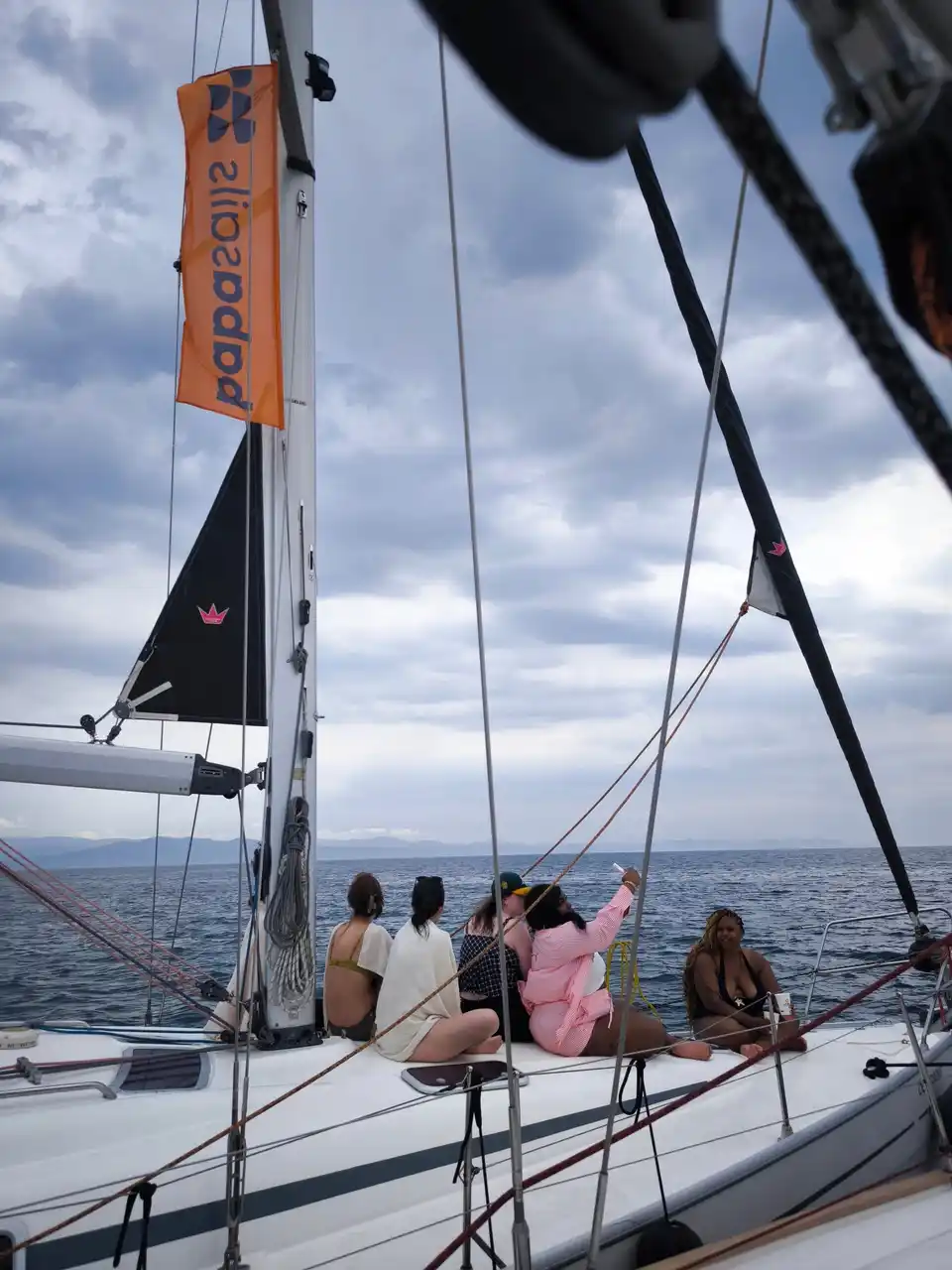 Women sitting on a sailing yacht’s bow under a cloudy sky, a flag on the mast
