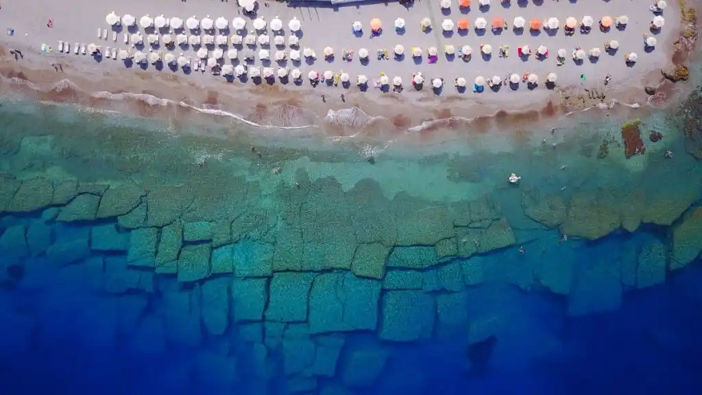 Beach umbrellas on the sand from above, flat rocks under clear water