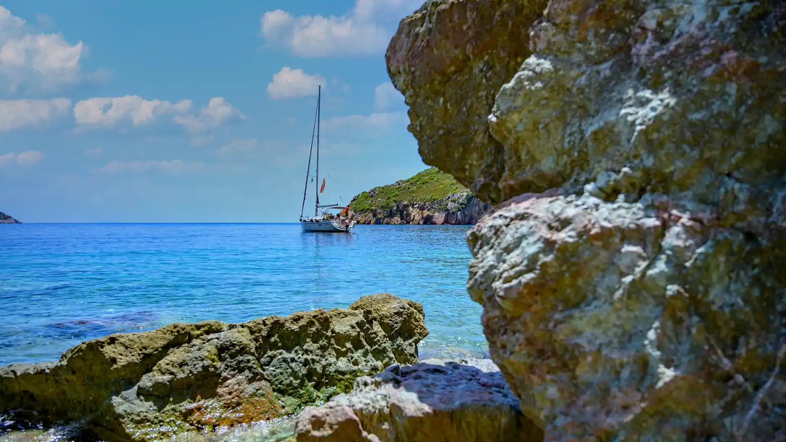 Sailing yacht anchored in a turquoise cove, seen past sunlit rocks