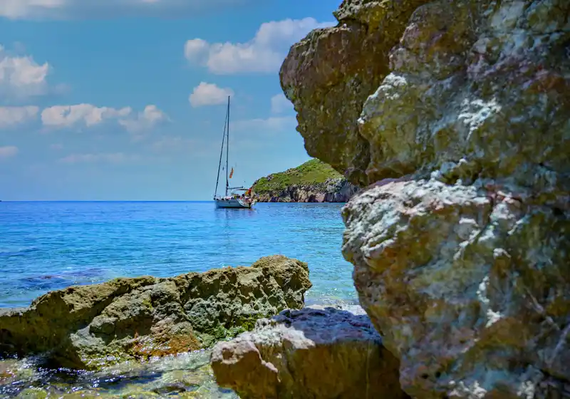 Sailing yacht anchored in a turquoise cove, seen past sunlit rocks