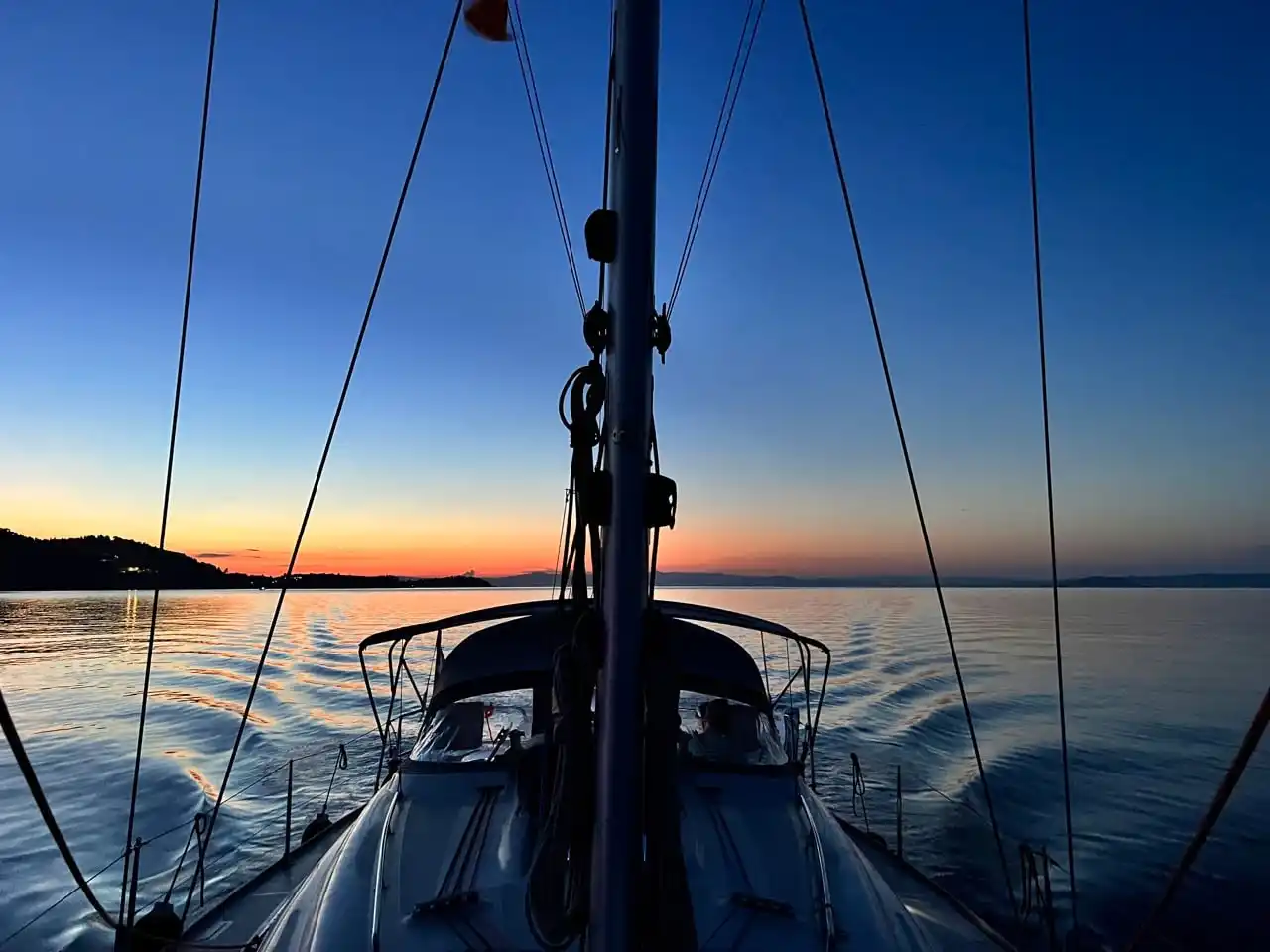 Calm sea at dusk seen from a sailing yacht’s bow, a glow on the horizon